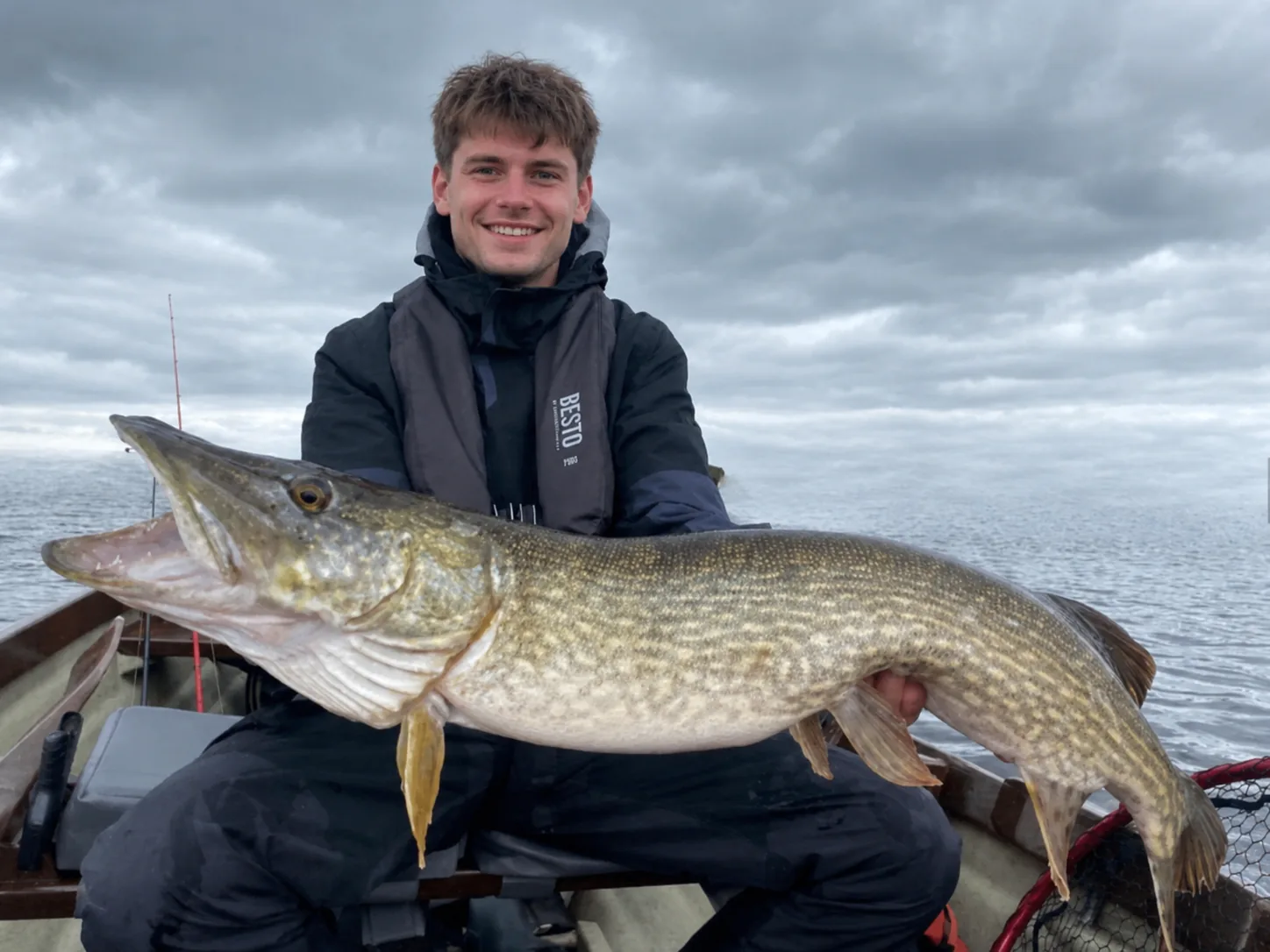 Angler holding a large pike on a boat in Ireland under cloudy skies.